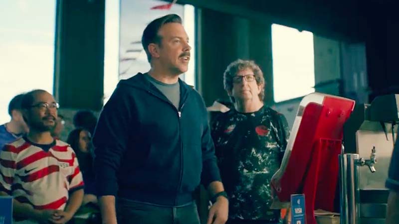 Jason Sudeikis ordering a hot dog at the stadium while other fans look on.