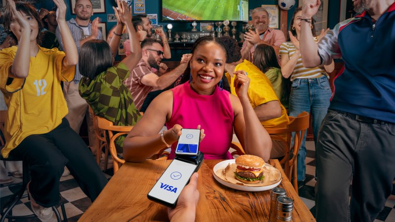Woman making contactless Visa payment at restaurant while football fans celebrate in the background.