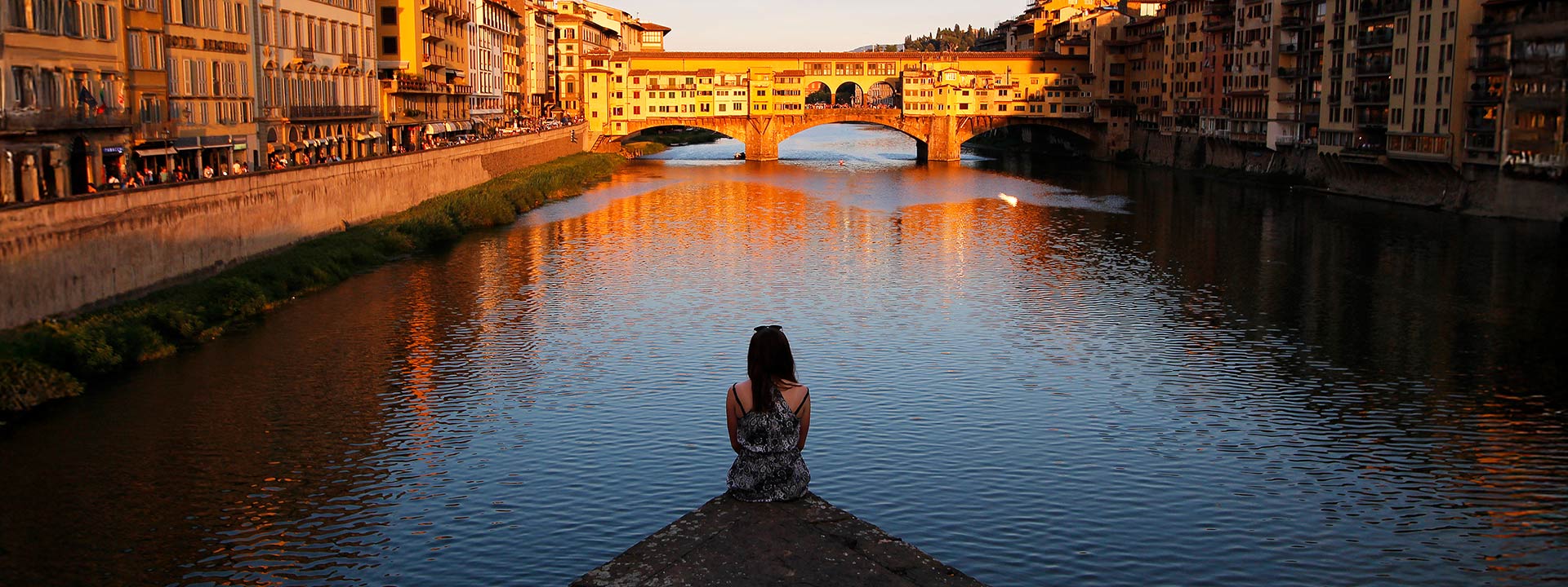 A person sits at the edge of a riverbank overlooking the Ponte Vecchio bridge in Florence, Italy, during sunset.