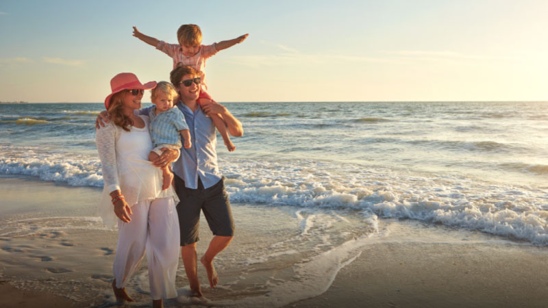 A family of four stands on the beach at sunset; two adults hold a toddler, while an older child sits on the man's shoulders with arms outstretched.