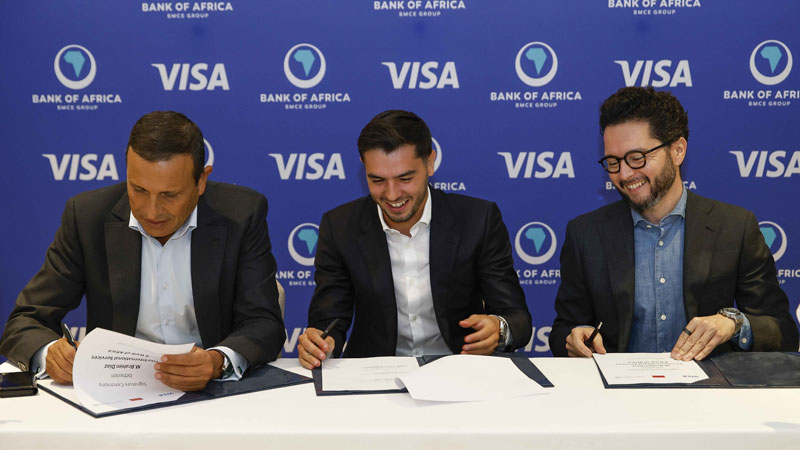 Three men in suits sign documents at a table, smiling, in front of a banner with Bank of Africa and Visa logos, conveying collaboration and positivity.