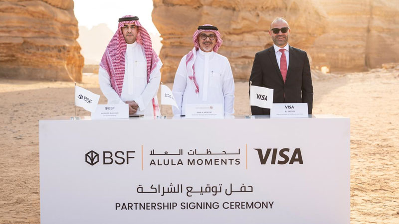 Three men stand behind a partnership signing ceremony table in a desert setting. The table displays logos for BSF, AlUla Moments, and Visa.