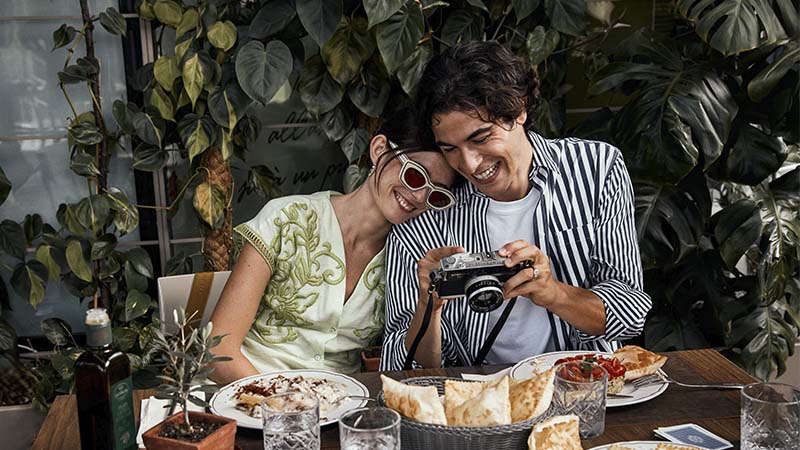 Two people sit at a table with food, smiling and looking at a camera together, surrounded by plants in the background.